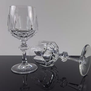 May include: Two clear crystal wine glasses featuring a faceted geometric pattern on the lower bowl and a multi-sided stem. One glass stands upright on a circular base, while the second glass lies horizontally, showcasing the intricate cut glass design.