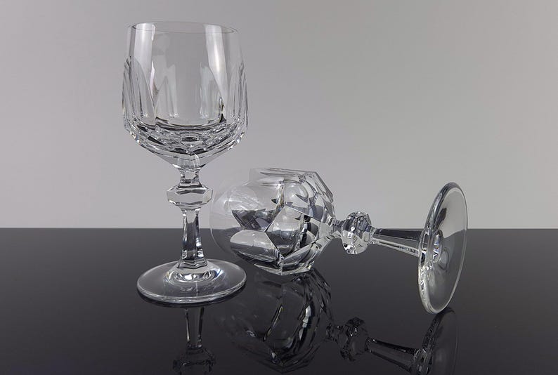 May include: Two clear crystal wine glasses featuring a faceted geometric pattern on the lower bowl and a multi-sided stem. One glass stands upright on a circular base, while the second glass lies horizontally, showcasing the intricate cut glass design.