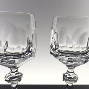 May include: Two clear crystal wine glasses featuring a faceted geometric bowl design with a wide, angular base and a multi-faceted stem. The glass surface displays sharp, clean lines and reflective edges that create a distinct, structured appearance.