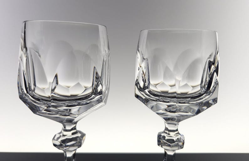 May include: Two clear crystal wine glasses featuring a faceted geometric bowl design with a wide, angular base and a multi-faceted stem. The glass surface displays sharp, clean lines and reflective edges that create a distinct, structured appearance.