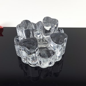 May include: A clear glass candle holder featuring a cluster of five heart-shaped indentations arranged in a circular pattern. The thick, textured glass has a heavy, solid appearance with smooth, rounded edges and a reflective, transparent surface.
