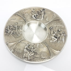 Peut inclure: Une assiette en métal argenté avec quatre scènes en relief représentant des femmes dans différentes poses. L'assiette a un puits central et une surface lisse et réfléchissante.