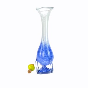 Puede incluir: Un jarrón alto de cristal transparente con la parte superior acampanada y un diseño azul moteado. El jarrón tiene una base bulbosa y un cuello delgado. Una pequeña manzana verde y un corcho están junto al jarrón.
