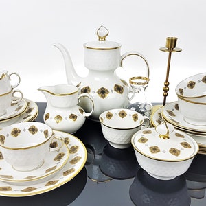 May include: A white porcelain tea set with gold trim and a textured surface. The set includes a teapot, creamer, sugar bowl, cups, saucers, and plates. Each piece is decorated with a brown floral pattern. A brass candlestick and a small glass vase are also present.
