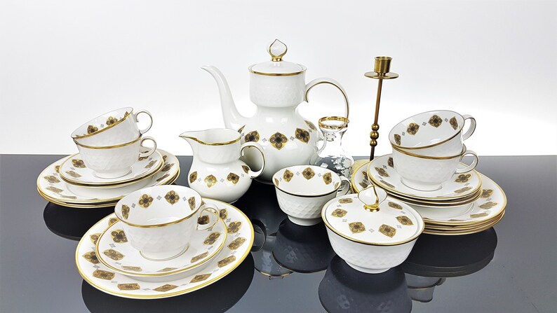 May include: A white porcelain tea set with gold trim and a textured surface. The set includes a teapot, creamer, sugar bowl, cups, saucers, and plates. Each piece is decorated with a brown floral pattern. A brass candlestick and a small glass vase are also present.