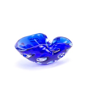 May include: A blue glass ashtray with a unique, organic shape. The ashtray has a textured surface and is made of thick glass.