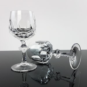 May include: Two clear crystal wine glasses featuring a faceted geometric bowl design. One glass stands upright on a circular base, while the second glass lies horizontally on its side. Both glasses have a multi-faceted stem and a smooth, round foot.