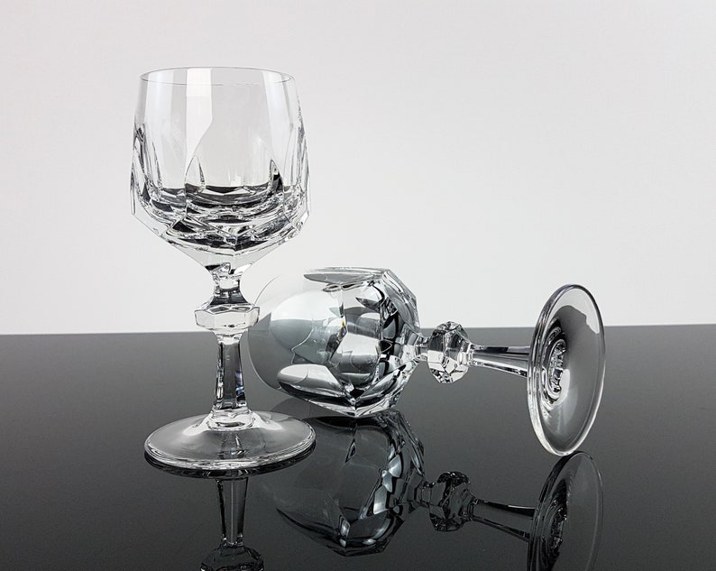 May include: Two clear crystal wine glasses featuring a faceted geometric bowl design. One glass stands upright on a circular base, while the second glass lies horizontally on its side. Both glasses have a multi-faceted stem and a smooth, round foot.