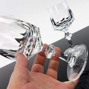 May include: Two clear crystal wine glasses featuring a faceted, geometric design on the bowl and a multi-sided stem. The glasses have a smooth, circular base and a reflective, transparent surface that catches light, creating sharp, angular shadows.