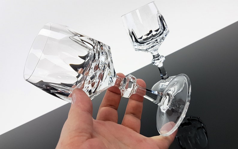 May include: Two clear crystal wine glasses featuring a faceted, geometric design on the bowl and a multi-sided stem. The glasses have a smooth, circular base and a reflective, transparent surface that catches light, creating sharp, angular shadows.