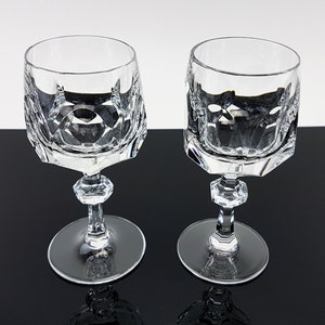 May include: Two clear crystal wine glasses featuring a faceted geometric pattern on the bowl, a hexagonal stem with a central decorative knob, and a smooth circular base. The glasses are transparent with sharp, angular cut glass detailing.