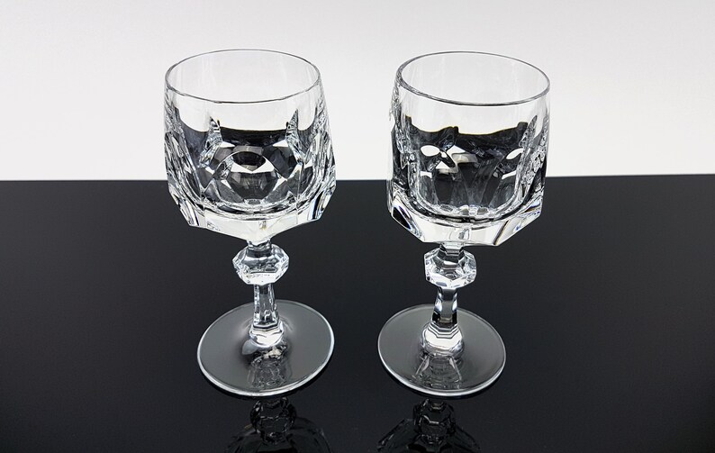 May include: Two clear crystal wine glasses featuring a faceted geometric pattern on the bowl, a hexagonal stem with a central decorative knob, and a smooth circular base. The glasses are transparent with sharp, angular cut glass detailing.