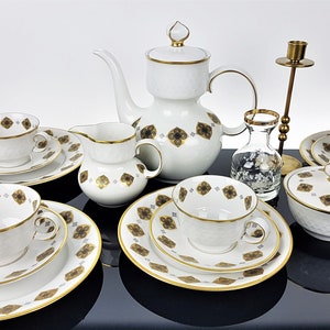 May include: A white porcelain tea set with gold trim and a repeating brown floral pattern. The set includes cups, saucers, plates, a teapot, creamer, sugar bowl, and a small vase. A brass candlestick holder is also present.