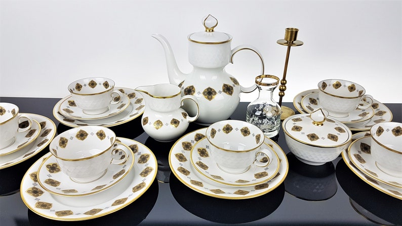 May include: A white porcelain tea set with gold trim and a repeating brown floral pattern. The set includes cups, saucers, plates, a teapot, creamer, sugar bowl, and a small vase. A brass candlestick holder is also present.