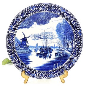 Könnte beinhalten: Blau-weiße Zierteller mit einer Landschaftsdarstellung mit Windmühle, Boot und Bäumen. Der Teller hat ein detailliertes Bordürenmuster. Der Teller wird auf einem goldfarbenen Ständer präsentiert.