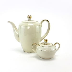 Może przedstawiać: Biały porcelanowy dzbanek do kawy i cukiernica z pozłacanymi zdobieniami. Dzbanek ma zaokrąglone ciało i dzióbek po boku. Cukiernica ma pokrywkę i uchwyt.