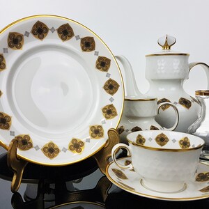 May include: A white porcelain tea set with gold trim and a repeating pattern of brown and gold floral designs. The set includes a plate, teapot, teacup, saucer, and a candlestick holder. The plate is approximately 10 inches in diameter.