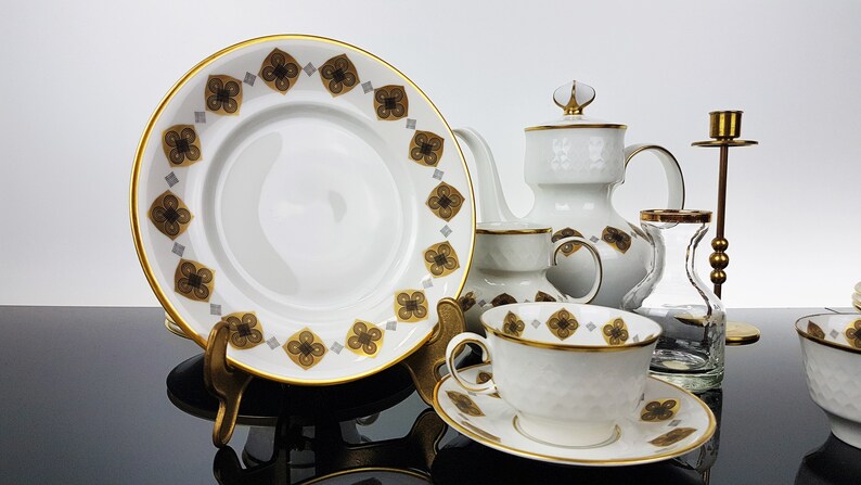 May include: A white porcelain tea set with gold trim and a repeating pattern of brown and gold floral designs. The set includes a plate, teapot, teacup, saucer, and a candlestick holder. The plate is approximately 10 inches in diameter.