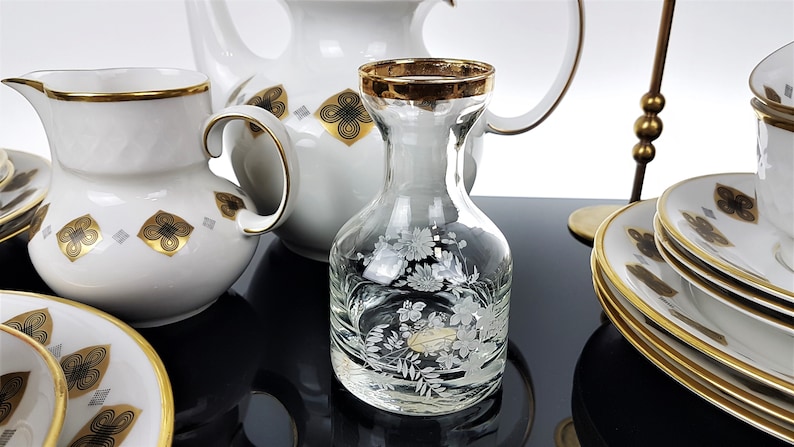 May include: A collection of white porcelain tableware with gold trim and decorative floral and geometric patterns. Includes a pitcher, creamer, plates, and a clear glass carafe with etched floral designs.
