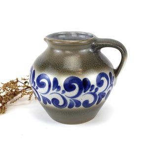 Könnte beinhalten: Kleiner, grauer Keramikkrug mit Henkel und Ausguss. Der Krug hat ein blau-weißes Blumendesign um den Körper. Der Krug wird wahrscheinlich zum Servieren von Flüssigkeiten verwendet.