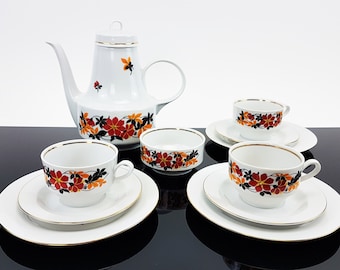 Könnte beinhalten: Ein weißes Porzellan-Tee-Set mit einem floralen Muster in Rot-, Orange- und Schwarztönen. Das Set beinhaltet eine Teekanne, eine Zuckerdose, eine Milchkännchen und vier Teetassen mit Untertassen.
