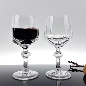 May include: Two clear crystal wine glasses with a faceted geometric bowl design and a multi-sided stem. One glass is filled with dark red wine, while the other is empty. The glasses feature a wide circular base and a distinct angular cut pattern.