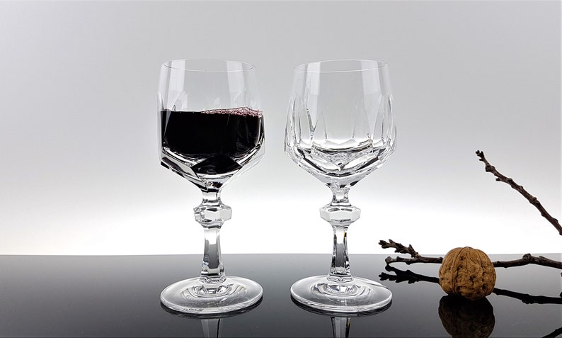 May include: Two clear crystal wine glasses with a faceted geometric bowl design and a multi-sided stem. One glass is filled with dark red wine, while the other is empty. The glasses feature a wide circular base and a distinct angular cut pattern.