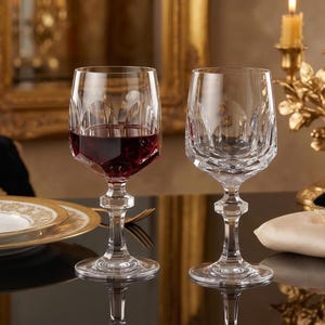 May include: Two clear crystal wine glasses with a faceted geometric pattern on the bowl and a multi-faceted stem. One glass is filled with red wine, while the other is empty. The glasses feature a wide base and a distinct, angular design.