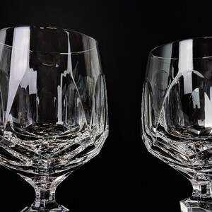May include: Two clear crystal glasses featuring a faceted geometric pattern on the lower bowl and a short, thick stem. The glasses have a wide, rounded rim and a heavy base, with light reflecting off the intricate cut glass surfaces.