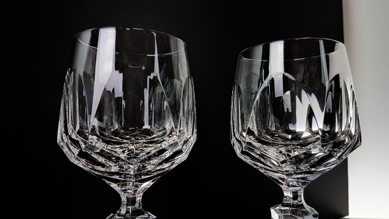 May include: Two clear crystal glasses featuring a faceted geometric pattern on the lower bowl and a short, thick stem. The glasses have a wide, rounded rim and a heavy base, with light reflecting off the intricate cut glass surfaces.