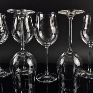 Könnte beinhalten: Eine Ansammlung von fünf Weingläsern aus klarem Glas mit schlanken Stielen und geschwungenen Kelchen. Drei Gläser stehen aufrecht auf ihren runden Füßen, während zwei Gläser umgedreht auf ihren Rändern stehen, um ihre Struktur zu zeigen.