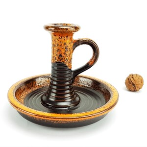 Peut inclure: Un bougeoir en céramique brun et orange avec une poignée. Le bougeoir a un motif en spirale noir et orange. Le bougeoir est posé sur une surface blanche.