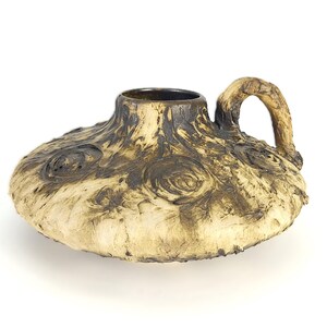Peut inclure: Un récipient en céramique à profil bas avec une seule anse, présentant une surface texturée de couleur crème avec des motifs organiques tourbillonnants brun foncé et un bord intérieur sombre. L'objet a une base large et plate.