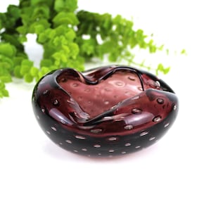 May include: A dark purple glass bowl with a textured surface and small white bubbles. The bowl has a heart-shaped opening. The bowl is on a white surface, with green foliage in the background.