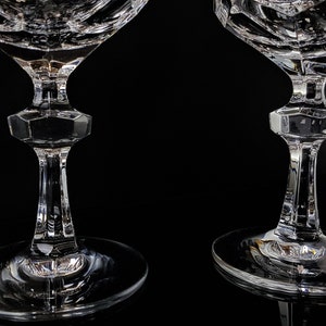 May include: Two clear crystal glasses featuring intricate geometric cut patterns on the bowls. Each glass has a faceted, hexagonal-shaped stem section that transitions into a smooth, circular base, showcasing reflective surfaces and sharp edges.