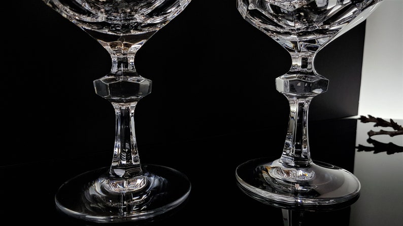 May include: Two clear crystal glasses featuring intricate geometric cut patterns on the bowls. Each glass has a faceted, hexagonal-shaped stem section that transitions into a smooth, circular base, showcasing reflective surfaces and sharp edges.