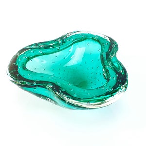 May include: A teal green glass ashtray with a bubbled texture. The ashtray is shaped like a heart and has a smooth, rounded edge.