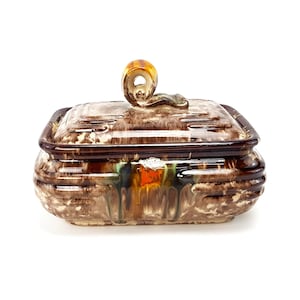 Könnte beinhalten: Eine quadratische Keramikdose mit Deckel. Die Dose ist mit brauner, weißer, grüner und orangefarbener Glasur verziert. Der Deckel hat einen einzigartigen, gerollten Griff.