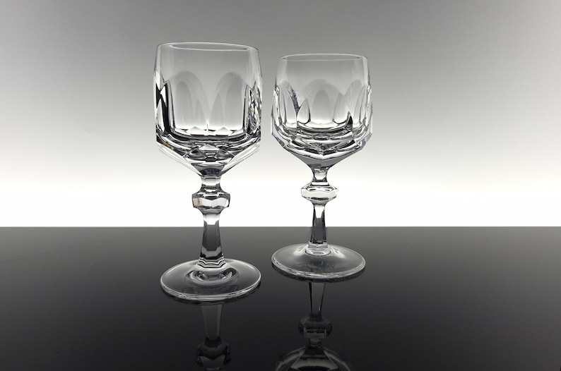 May include: Two clear crystal wine glasses featuring a faceted, geometric bowl design with vertical cut patterns. Each glass has a multi-faceted stem and a circular base, showcasing a reflective, transparent finish with sharp, angular edges.