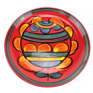 Könnte beinhalten: Ein roter Keramikteller mit grünem Rand und einem farbenfrohen Blumendesign. Die Blume ist in Orange-, Gelb-, Grün-, Blau- und Rottönen bemalt.