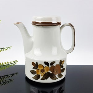 Puede incluir: Tetera de cerámica blanca con tapa marrón y diseño floral. La tetera tiene un asa marrón y una banda marrón alrededor de la tapa.