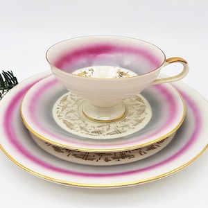 May include: A vintage teacup and saucer set with a pink and gray ombre design and gold accents. The set features a delicate floral pattern and a gold rim.