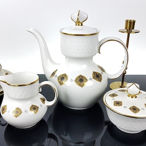 May include: A white porcelain tea set with gold trim and a geometric floral pattern. The set includes a teapot, creamer, sugar bowl, cups, saucers, and a small decorative glass bottle. A brass candlestick is also present.