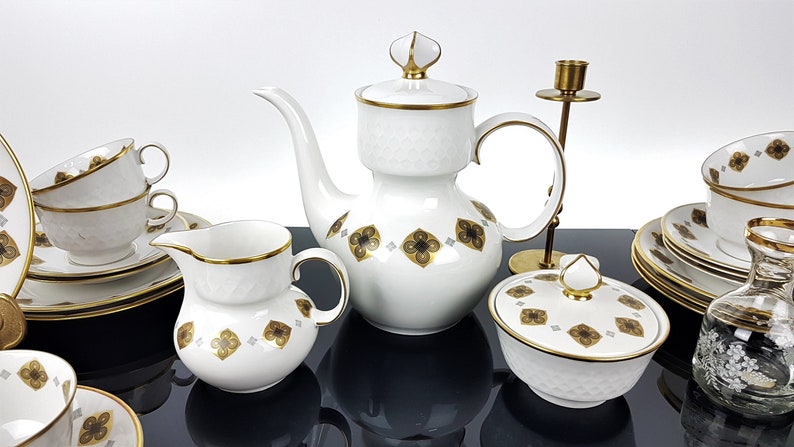 May include: A white porcelain tea set with gold trim and a geometric floral pattern. The set includes a teapot, creamer, sugar bowl, cups, saucers, and a small decorative glass bottle. A brass candlestick is also present.