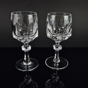 May include: Two clear crystal wine glasses featuring a faceted, geometric bowl design with a scalloped upper rim. Each glass has a multi-faceted stem and a circular base, showcasing a reflective, transparent quality throughout the structure.