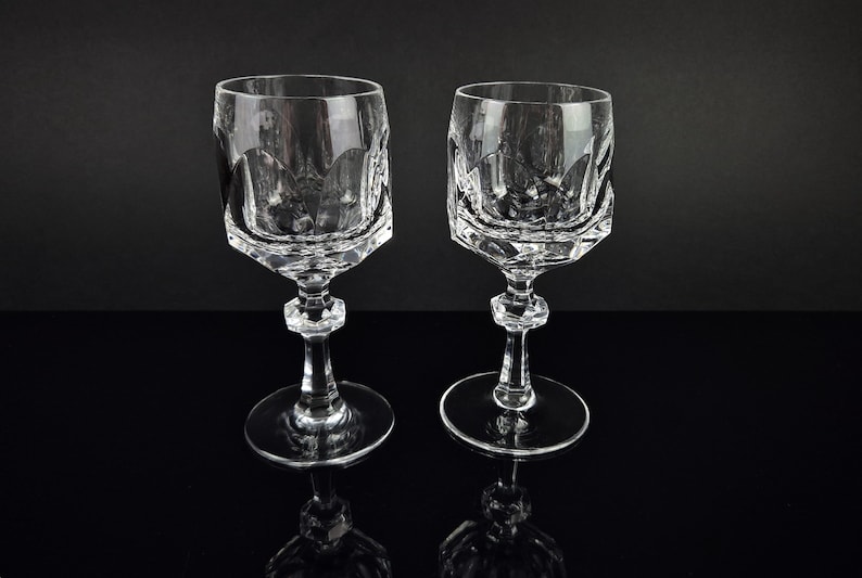 May include: Two clear crystal wine glasses featuring a faceted, geometric bowl design with a scalloped upper rim. Each glass has a multi-faceted stem and a circular base, showcasing a reflective, transparent quality throughout the structure.