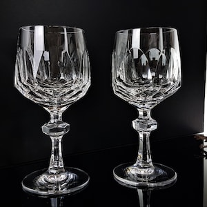 May include: Two clear crystal wine glasses featuring a faceted geometric pattern on the bowl and a hexagonal node on the stem. The glasses have a wide circular base and a smooth rim, showcasing intricate light-reflecting cuts throughout the glass.
