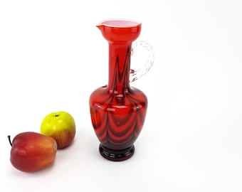 May include: A red glass vase with a white swirl design and a clear glass handle. The vase is sitting on a white surface with two red apples and one yellow apple.