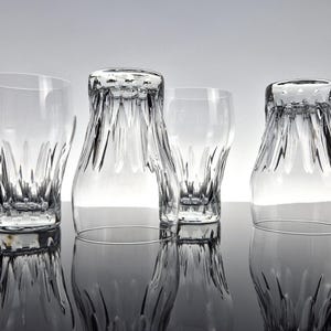 May include: Four clear, decorative drinking glasses with a textured, ridged design. Two glasses are upright, and two are inverted, creating a symmetrical arrangement. The glasses are reflected on a glossy surface.