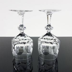 May include: Two clear crystal wine glasses positioned upside down. Each glass features a faceted, geometric bowl design and a multi-sectioned stem with a central hexagonal knob. The base of each glass is flat and circular, reflecting light clearly.